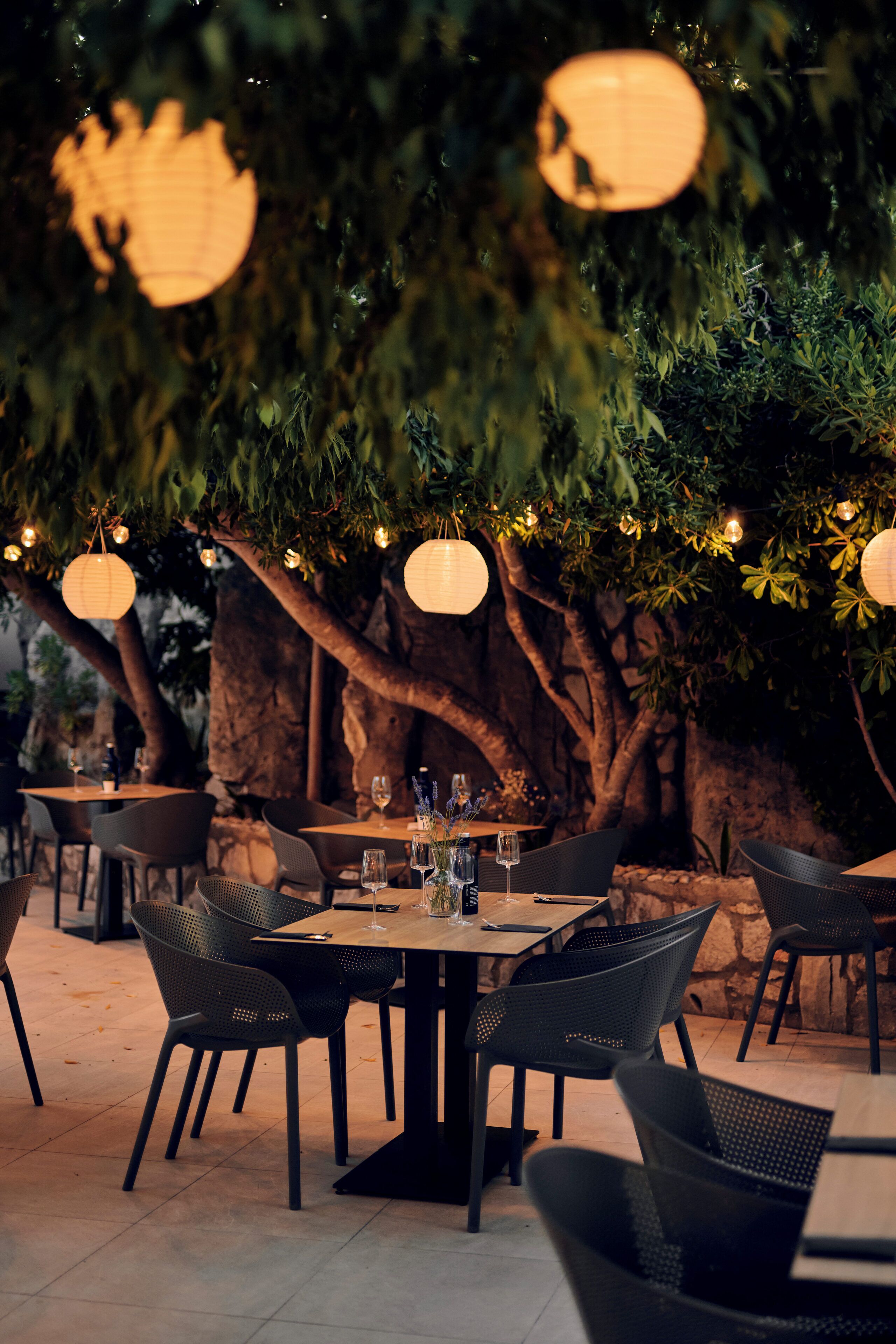 An outdoor dining area with tables and chairs, surrounded by trees and a stone wall.