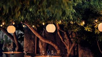 An outdoor dining area with tables and chairs, surrounded by trees and a stone wall.