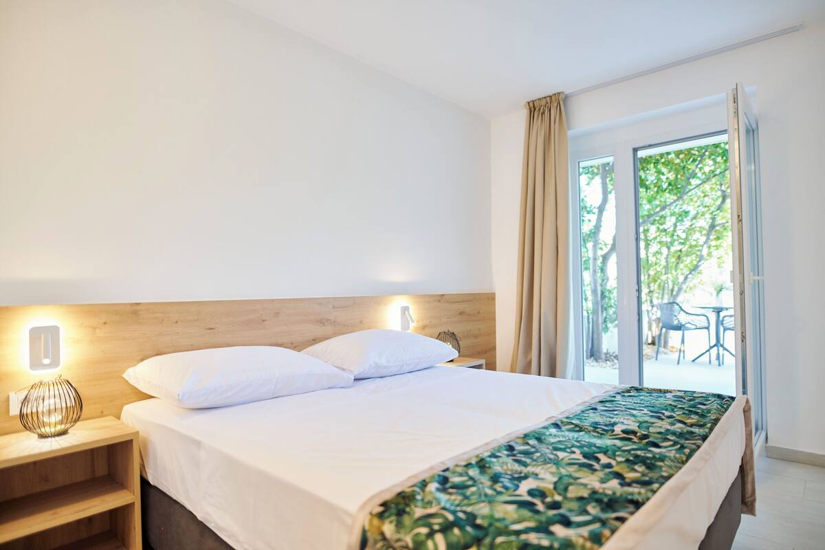 A hotel room with a bed, a nightstand, a lamp, and a view of the outdoors through a glass door.