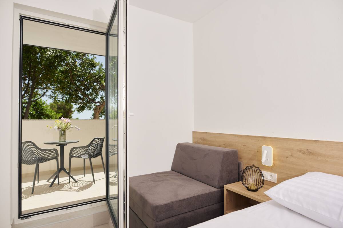 A modern hotel room with a sofa bed, a small bedside table with a lamp, and a view of an outdoor patio with a table and chairs.