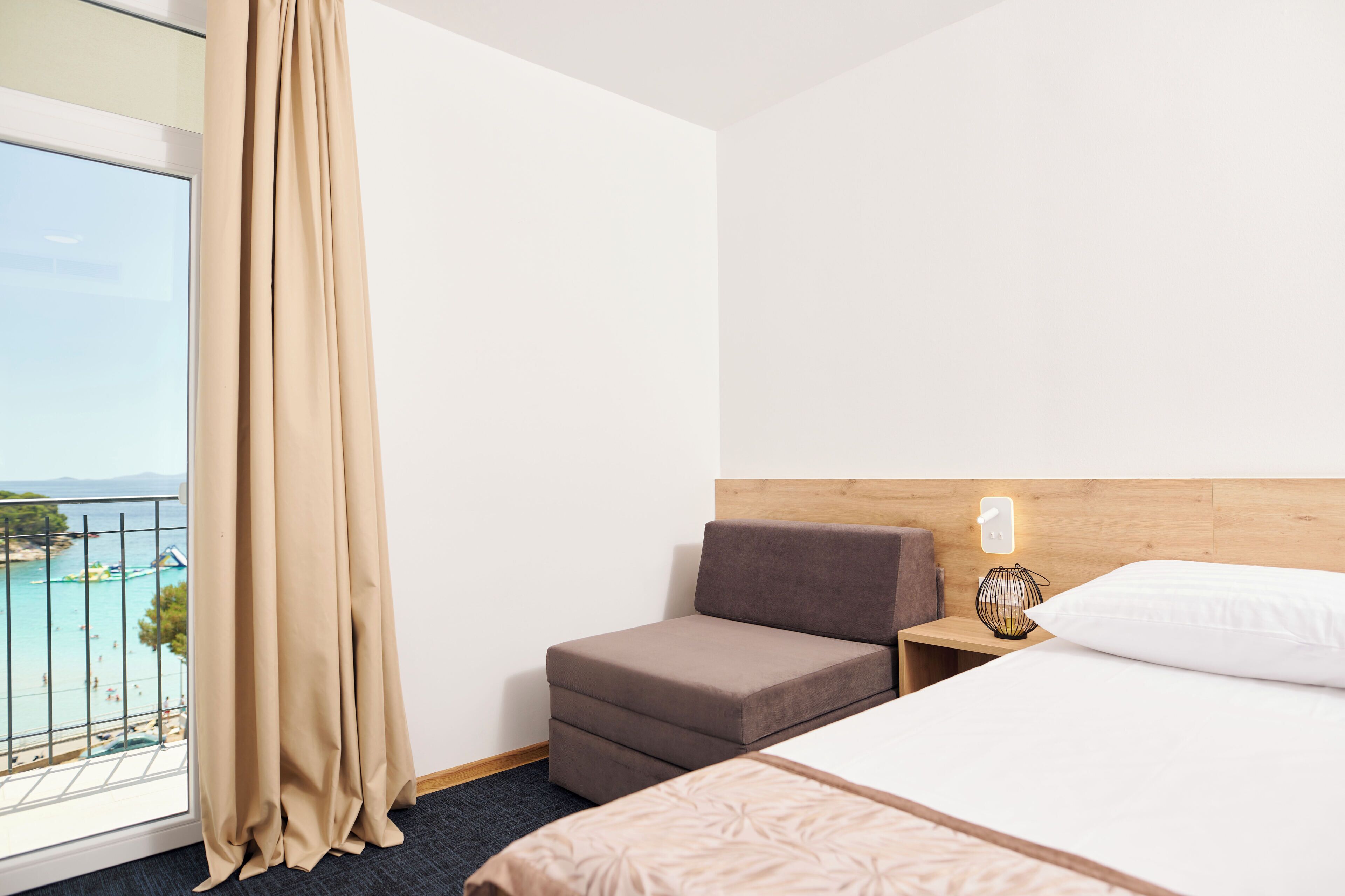 Habitación de hotel con cama, un pequeño sofá, mesita de noche, lámpara y vistas a la playa y la piscina.