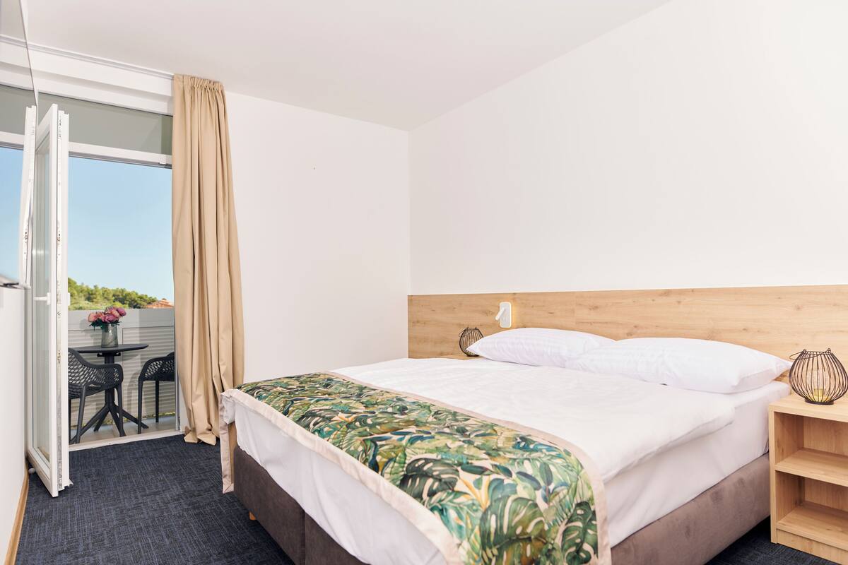 A hotel room with a bed, a wooden headboard, a nightstand, a table with chairs, and a view through a glass door.