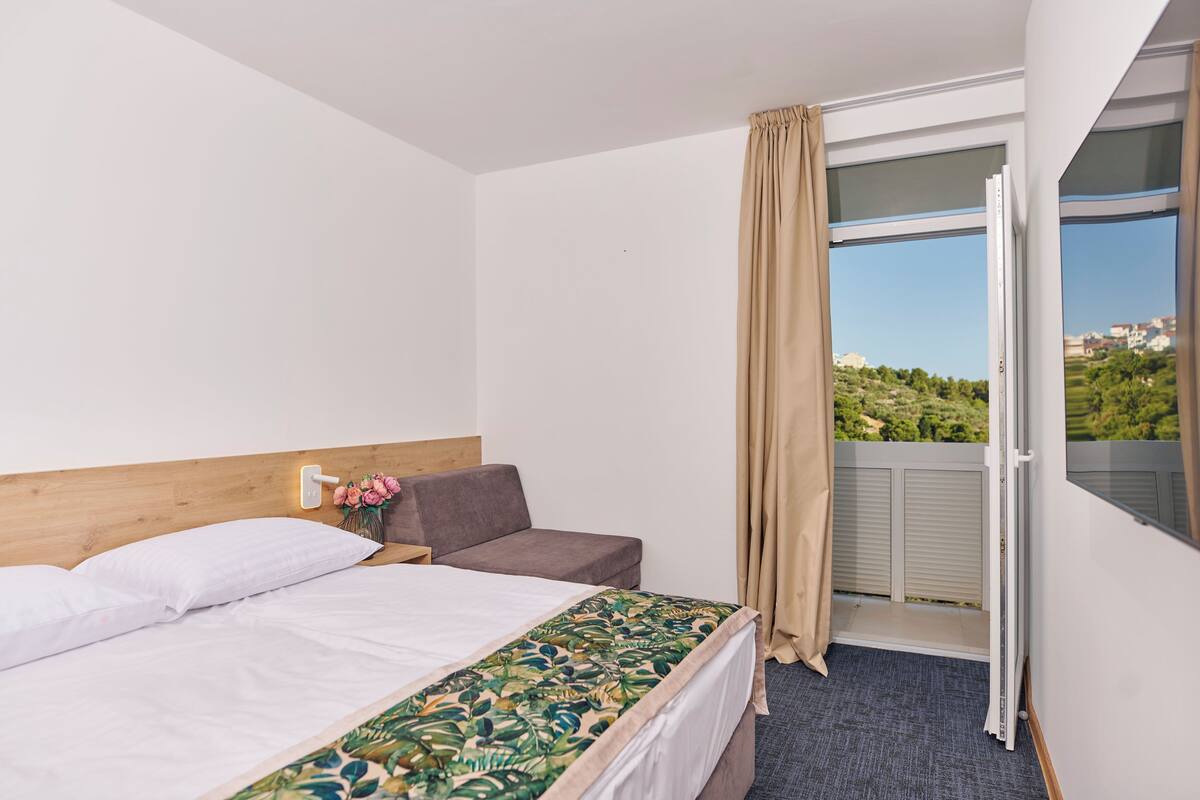 A hotel room with a bed, a small sofa, a nightstand, a window with a view of greenery, and a curtain.