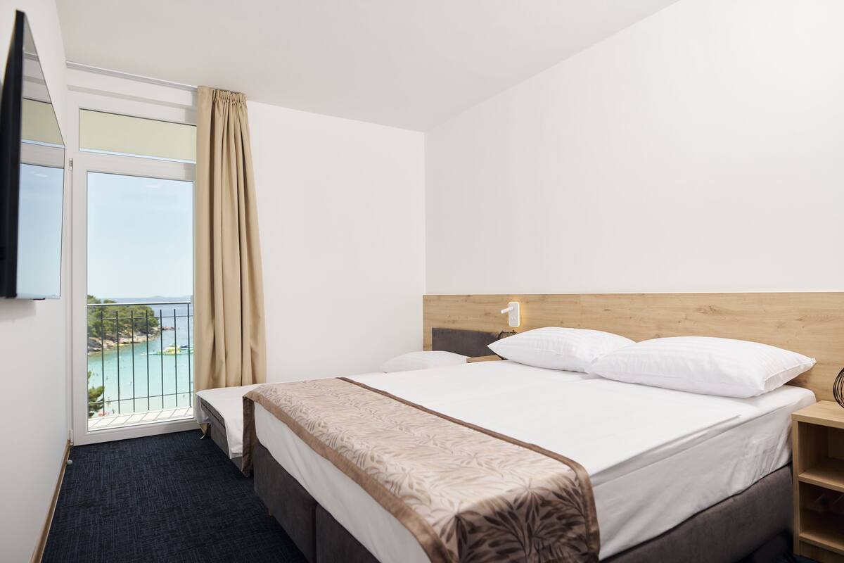 A hotel room with a bed, a bedside table, a TV, a window with a view of the sea, and a balcony.
