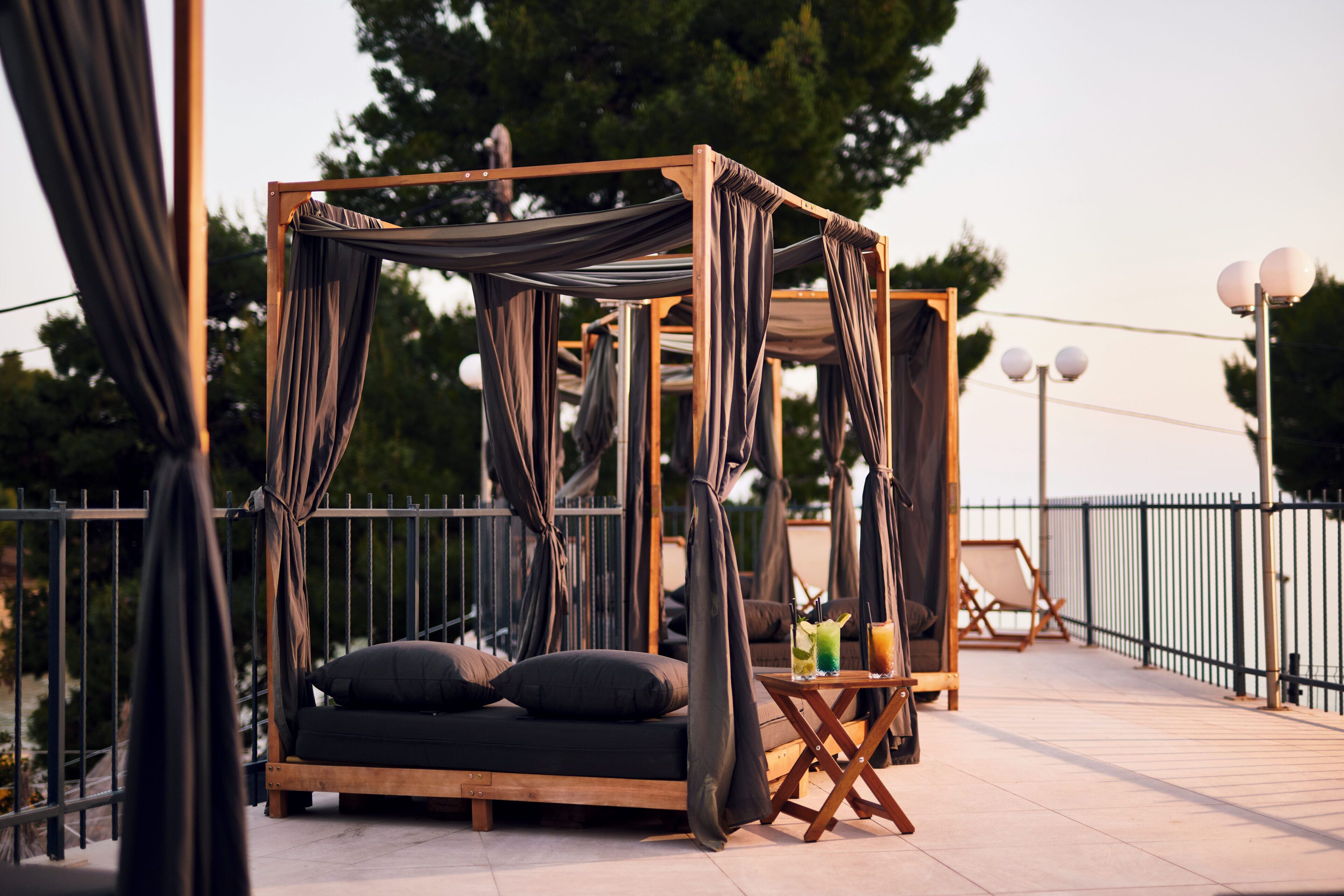Una cama con dosel de madera, cortinas oscuras, una mesita con bebidas y un sillón de descanso en una terraza al aire libre con barandilla y árboles al fondo.