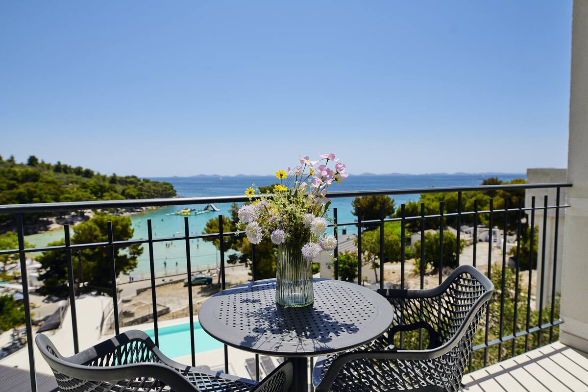 A balcony with a round table and chairs, a vase with flowers, and a view of a beach and sea.