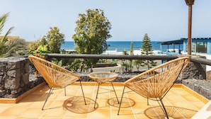 A terrace with wicker chairs and a small table, overlooking a beach and a building with a blue roof. - El Hotelito Del Golfo (Yaiza)