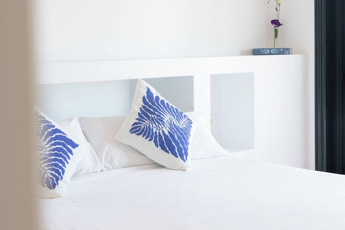 A neatly made bed with white bedding and blue patterned pillows, a small vase with a flower on the headboard, and a plain white wall.
