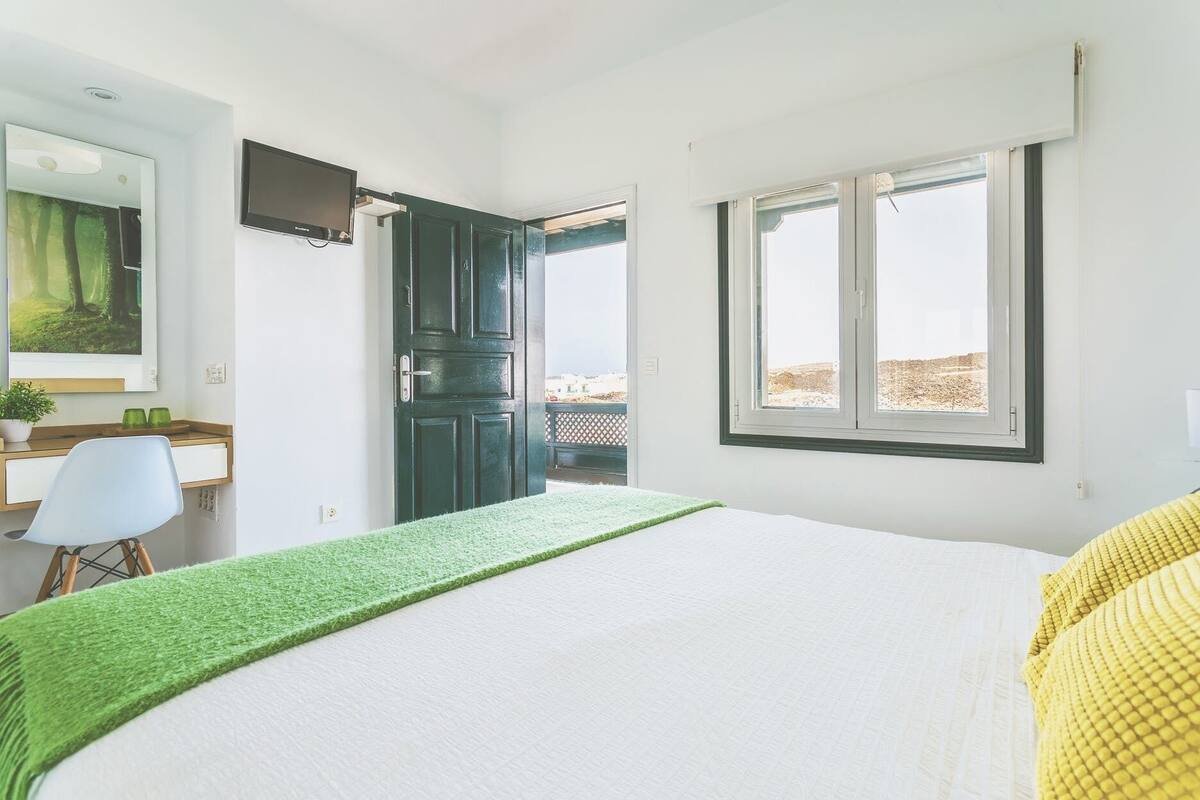 A bedroom with a bed, a desk, a chair, a window, and a door leading to an outdoor area.