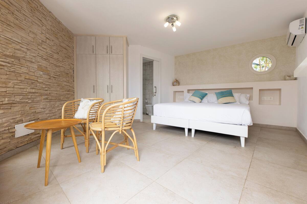 A bedroom with a bed, a wooden table, two wicker chairs, a stone wall, a ceiling light, and a window.