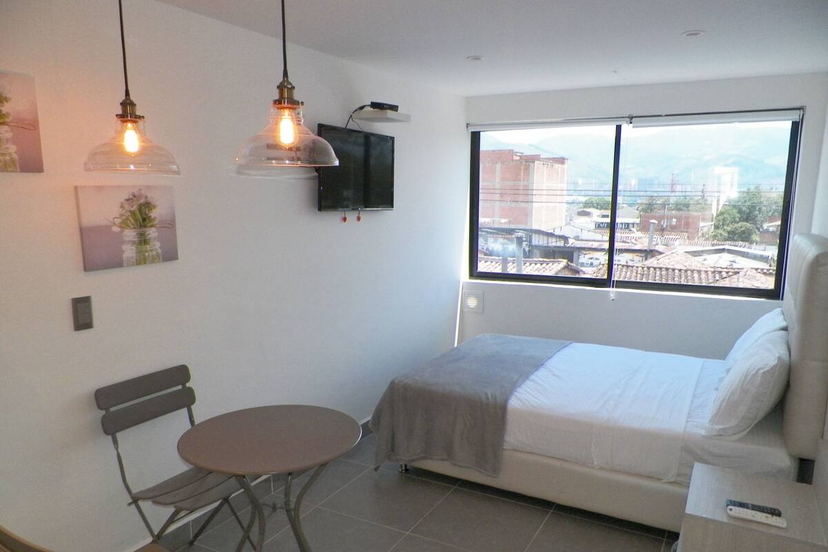 A bedroom with a bed, a small table, a bench, and a window with a city view.