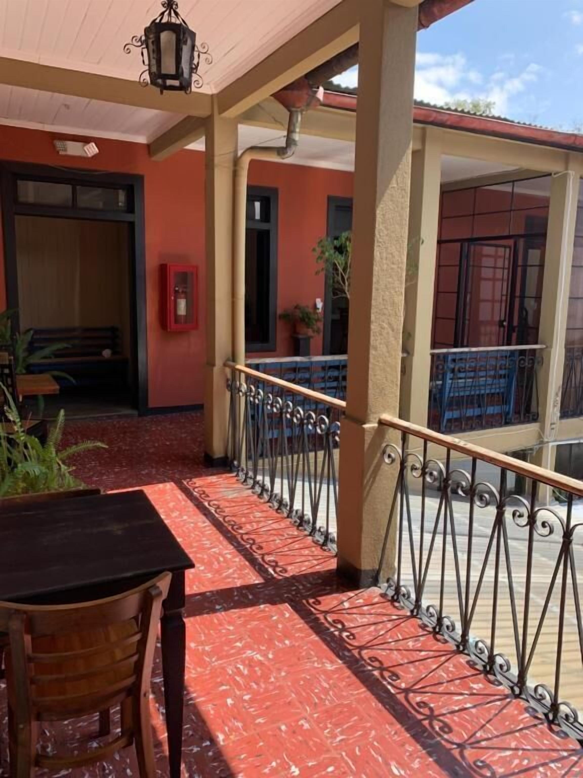 An outdoor patio area with a wooden table and chair, a red patterned floor, a metal railing, and a building with a red door and windows in the background.