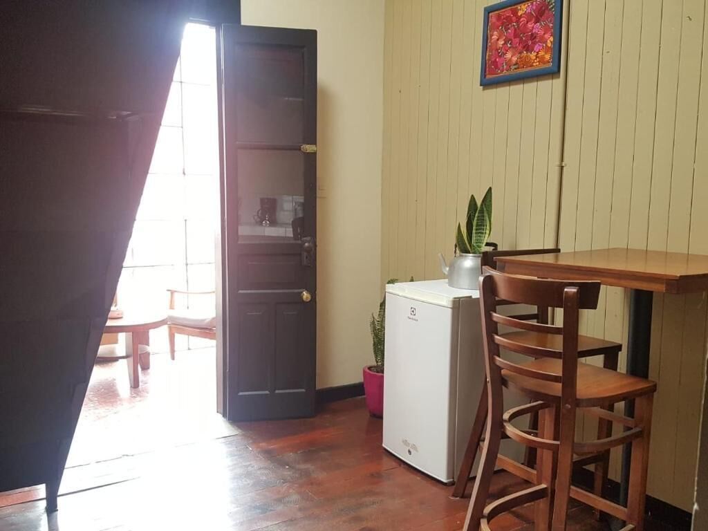 A room with a wooden table, a chair, a small refrigerator, a potted plant, and a door leading to another room.