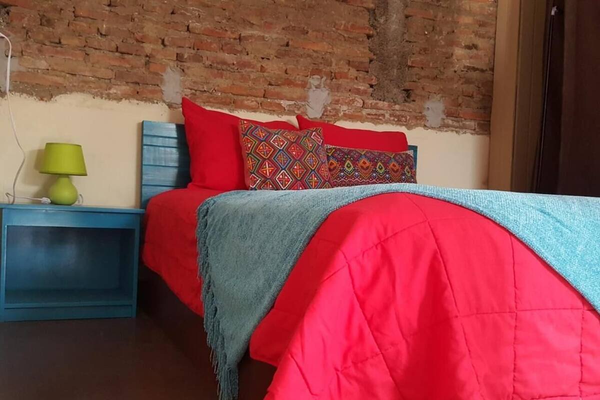 A bedroom with a brick wall, a bed with red bedding and patterned pillows, a blue nightstand with a green lamp, and a blue shelf.