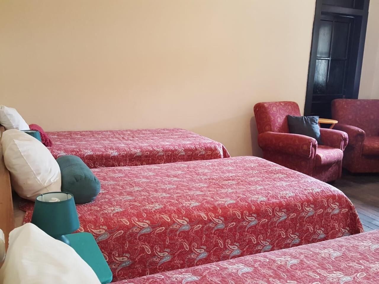 A room with two beds covered in red patterned bedspreads, a red armchair, and a small table with a lamp.