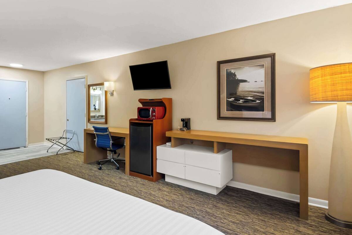 Quarto de hotel com cama, escrivaninha com computador, televisão, quadro emoldurado e luminária.