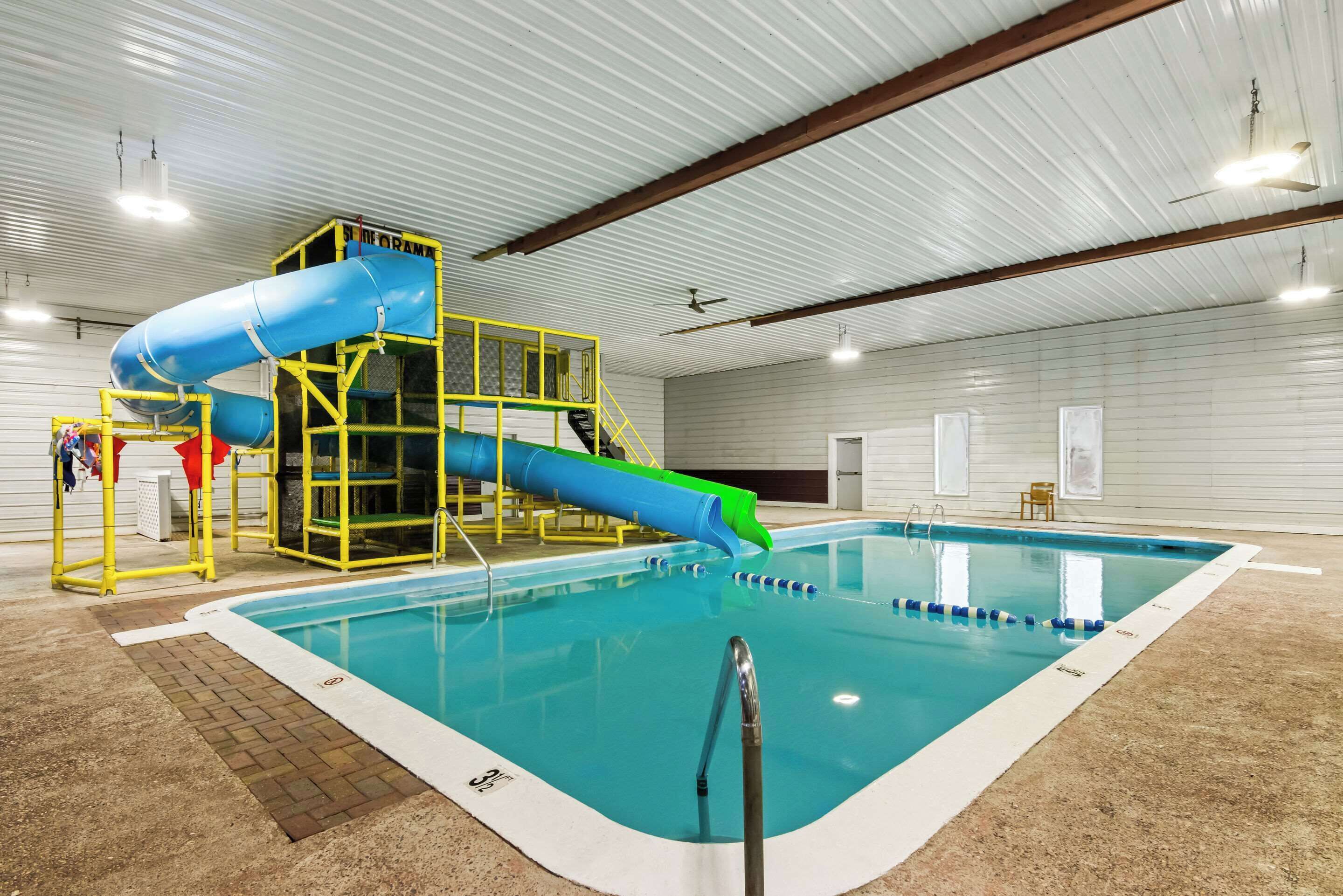 An indoor swimming pool with a blue and green slide, yellow safety railings, and a clear pool edge.