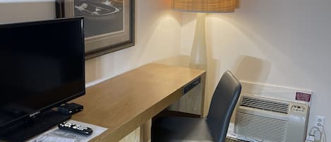 Habitación de hotel con un escritorio de madera, una silla de oficina negra, una lámpara de pie, un cuadro enmarcados con un paisaje y un aire acondicionado instalado en la pared.
