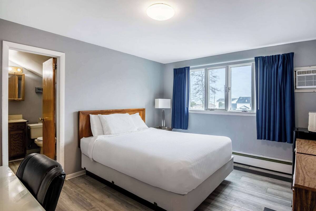 Quarto de hotel com cama, escrivaninha, cadeira, janela com cortinas e banheiro visível por uma porta aberta.