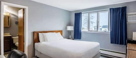 Une chambre d’hôtel avec un lit, un bureau, une chaise, une fenêtre avec des rideaux et une salle de bain visible à travers une porte ouverte.
