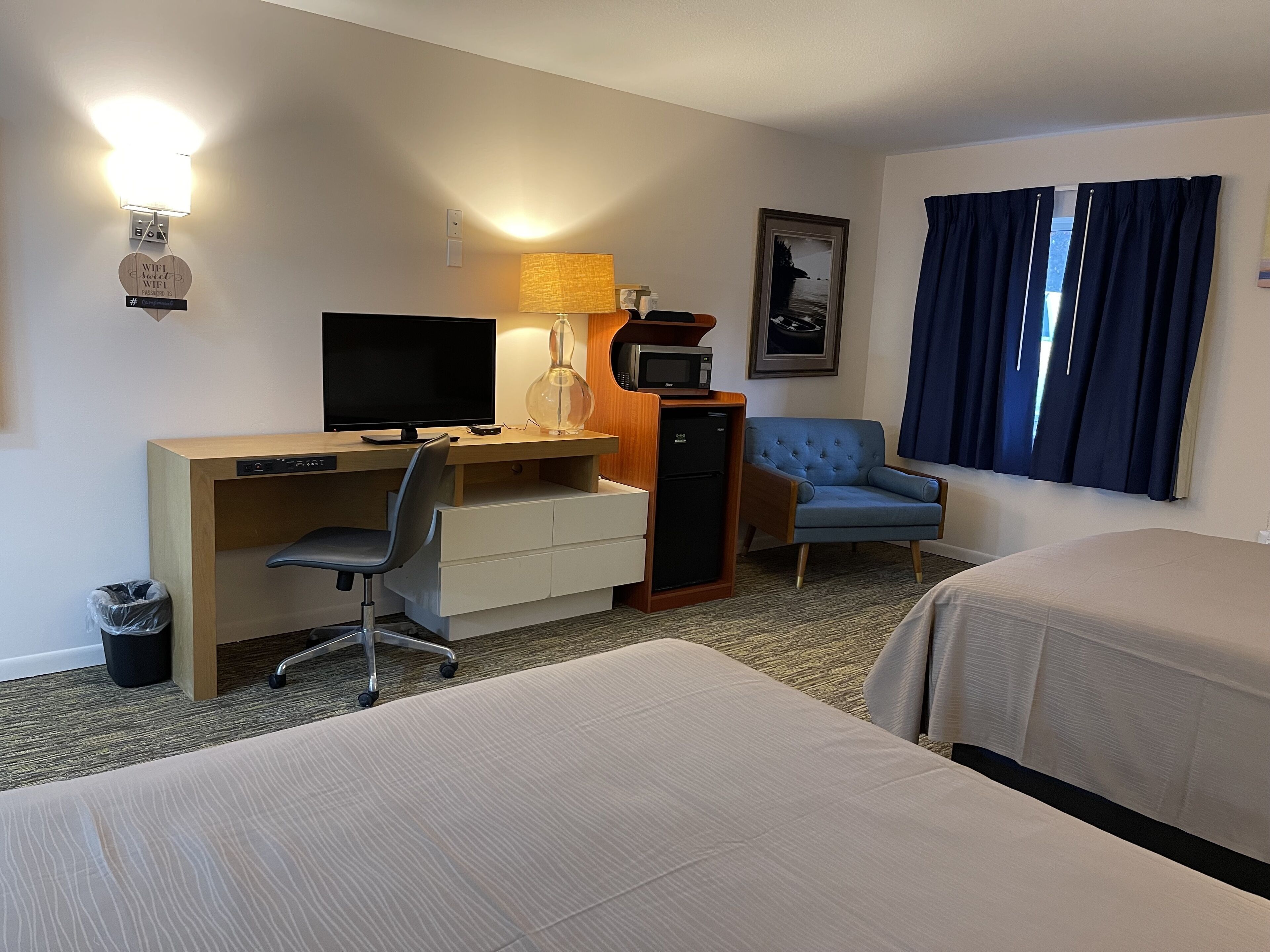 A hotel room with a desk, a television, a lamp, a chair, a bed, and a window with curtains.