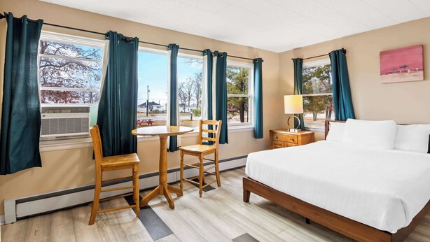 A hotel room with a bed, a desk with chairs, a lamp, and windows with curtains. - Rodeway Inn Oscoda (Oscoda)