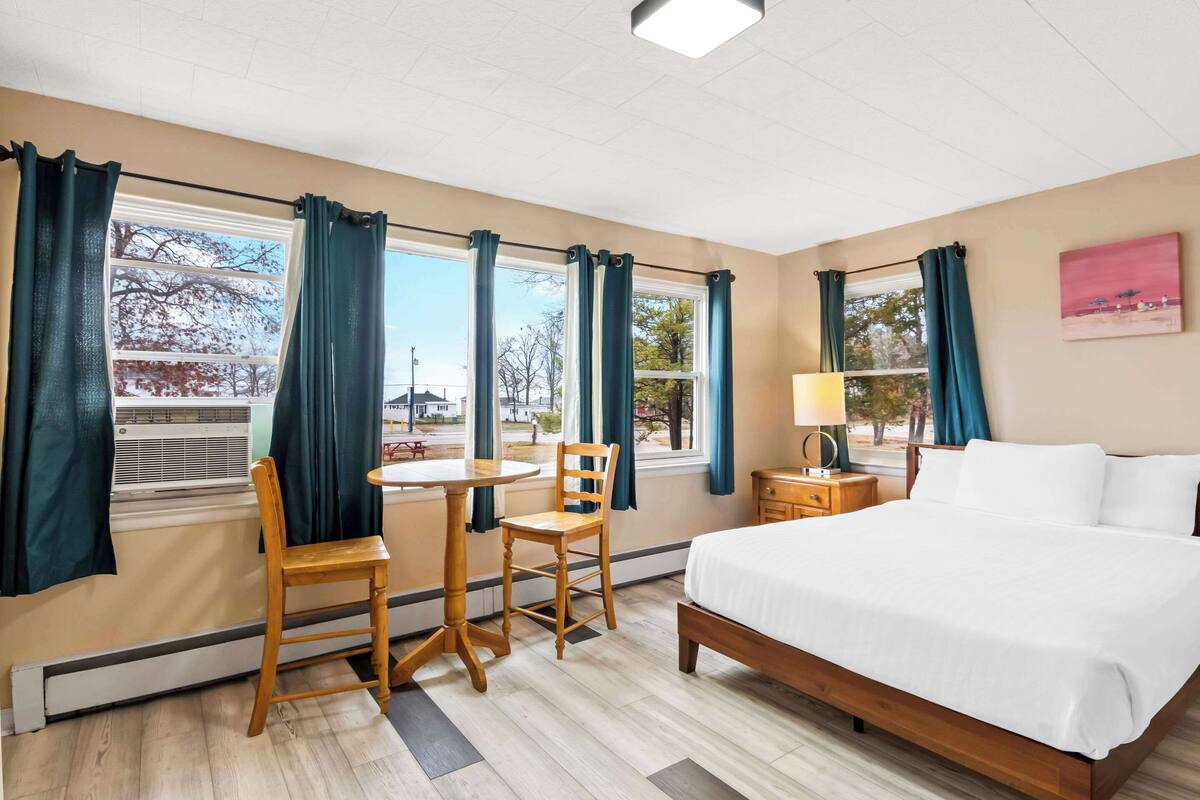 Quarto de hotel com cama, escrivaninha com cadeiras, luminária e janelas com cortinas.