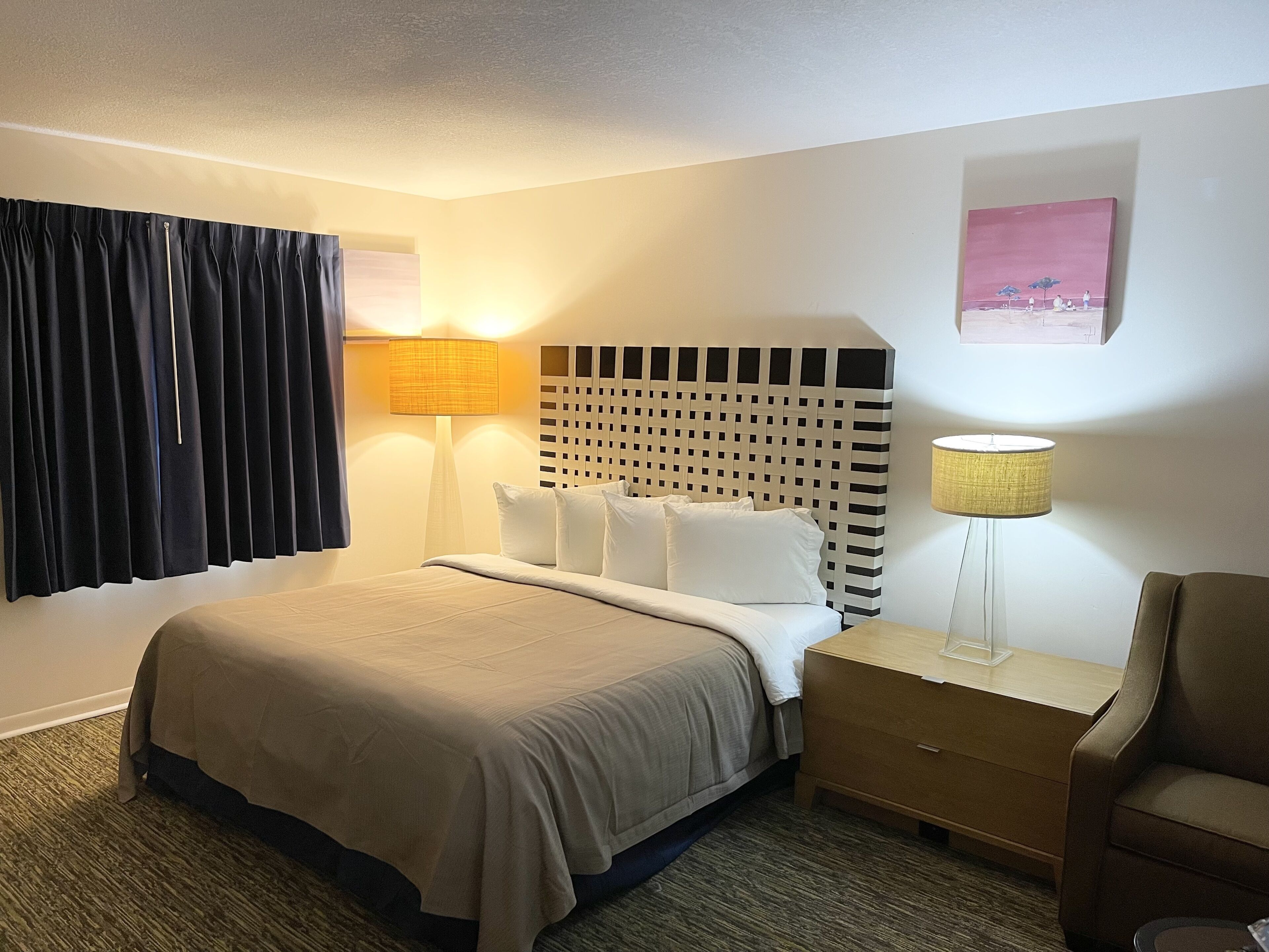 Una habitación de hotel con una cama, una mesita de noche, una silla, una lámpara y un cuadro en la pared.