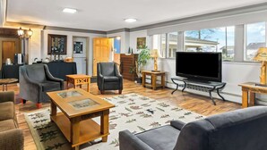 A spacious living room with a large TV, comfortable seating, a wooden coffee table, and a view through large windows. - Rodeway Inn Oscoda (Oscoda)