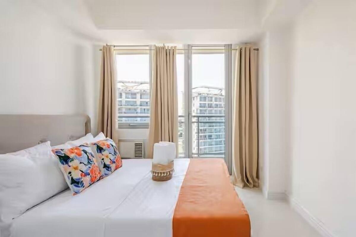A hotel room with a bed, a window with curtains, and a view of buildings outside.