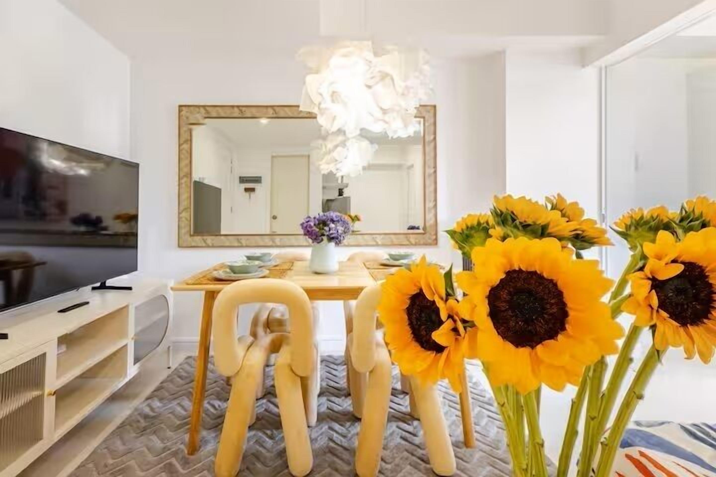 A modern living room with a television, a dining table with chairs, a large mirror, and sunflowers.