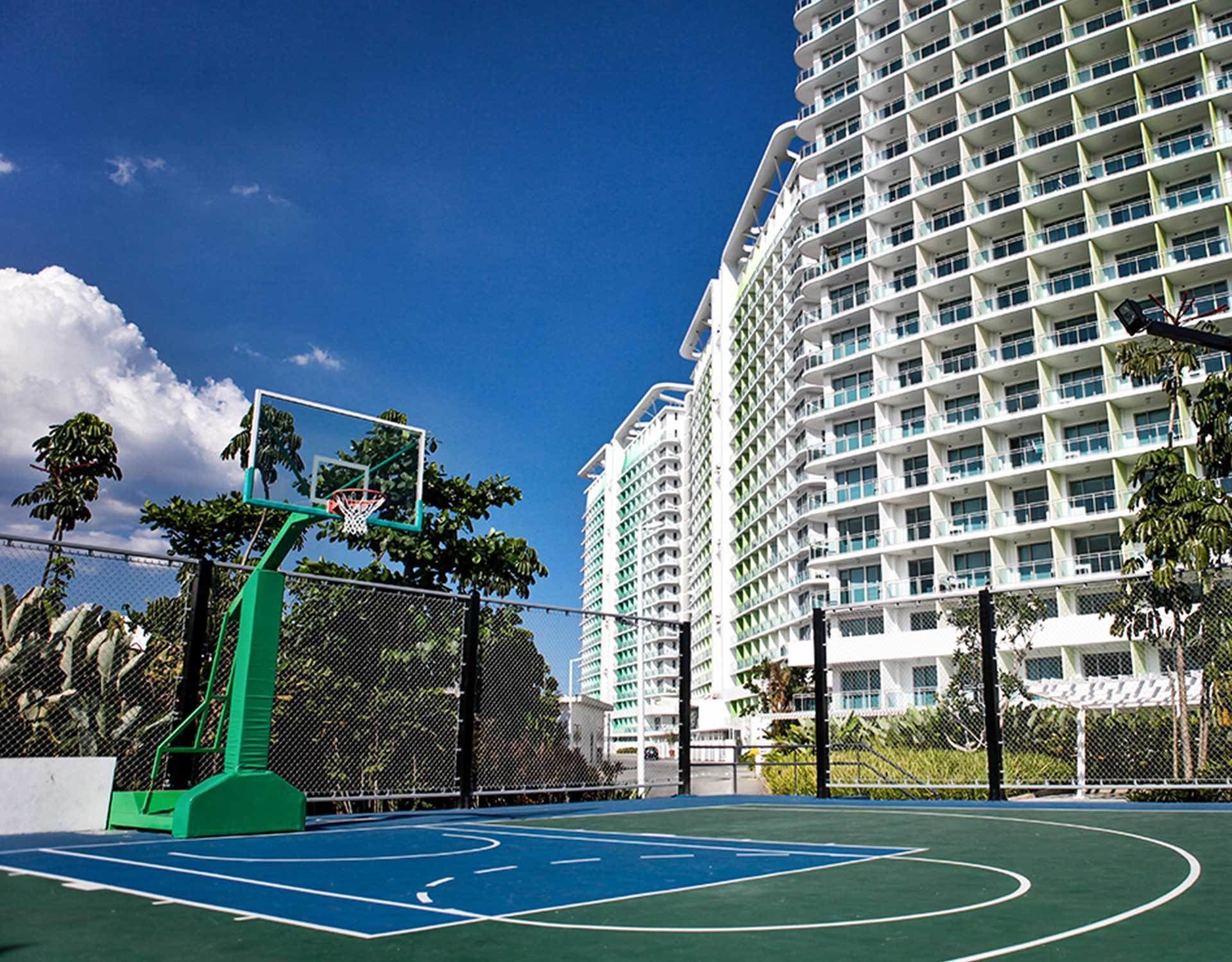 A basketball court with a green basketball hoop, surrounded by a chain-link fence and palm trees.