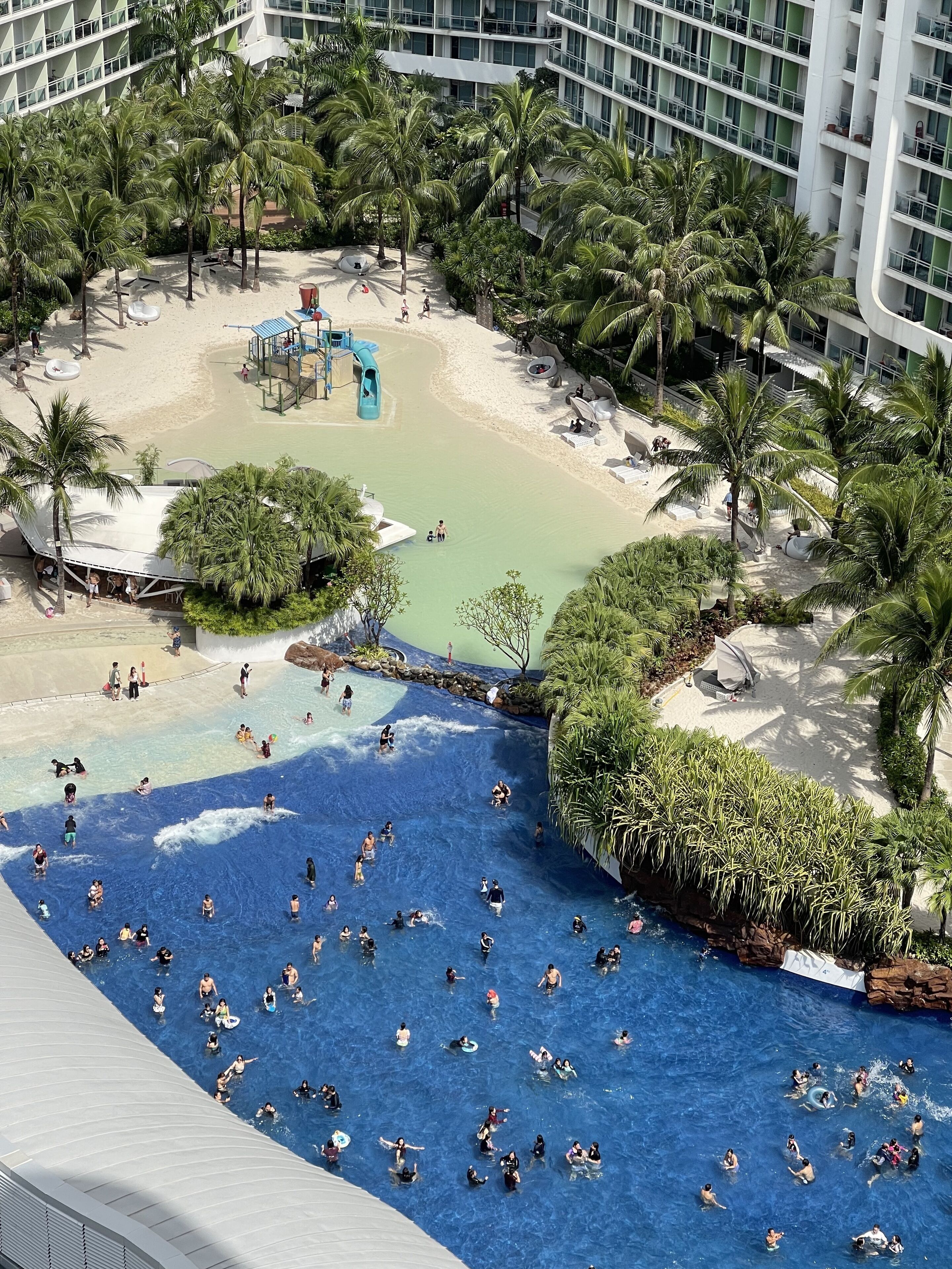 A resort with a large pool area, a sandy beach, and palm trees.