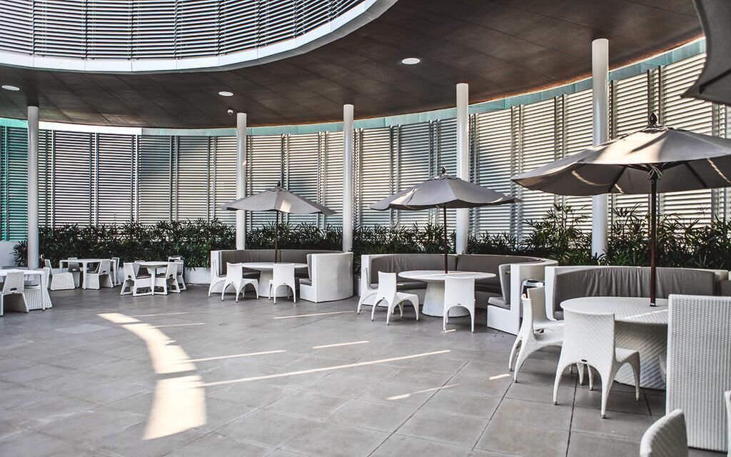 Un vaste espace extérieur aménagé avec des tables et des chaises blanches, entouré de verdure et doté de grandes fenêtres.