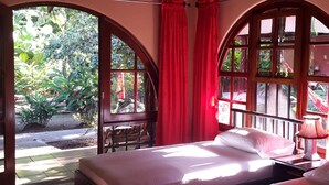 A room with a bed, a nightstand, and a window with red curtains. There is a view of greenery outside. - Paraíso Escondido (Jacó)