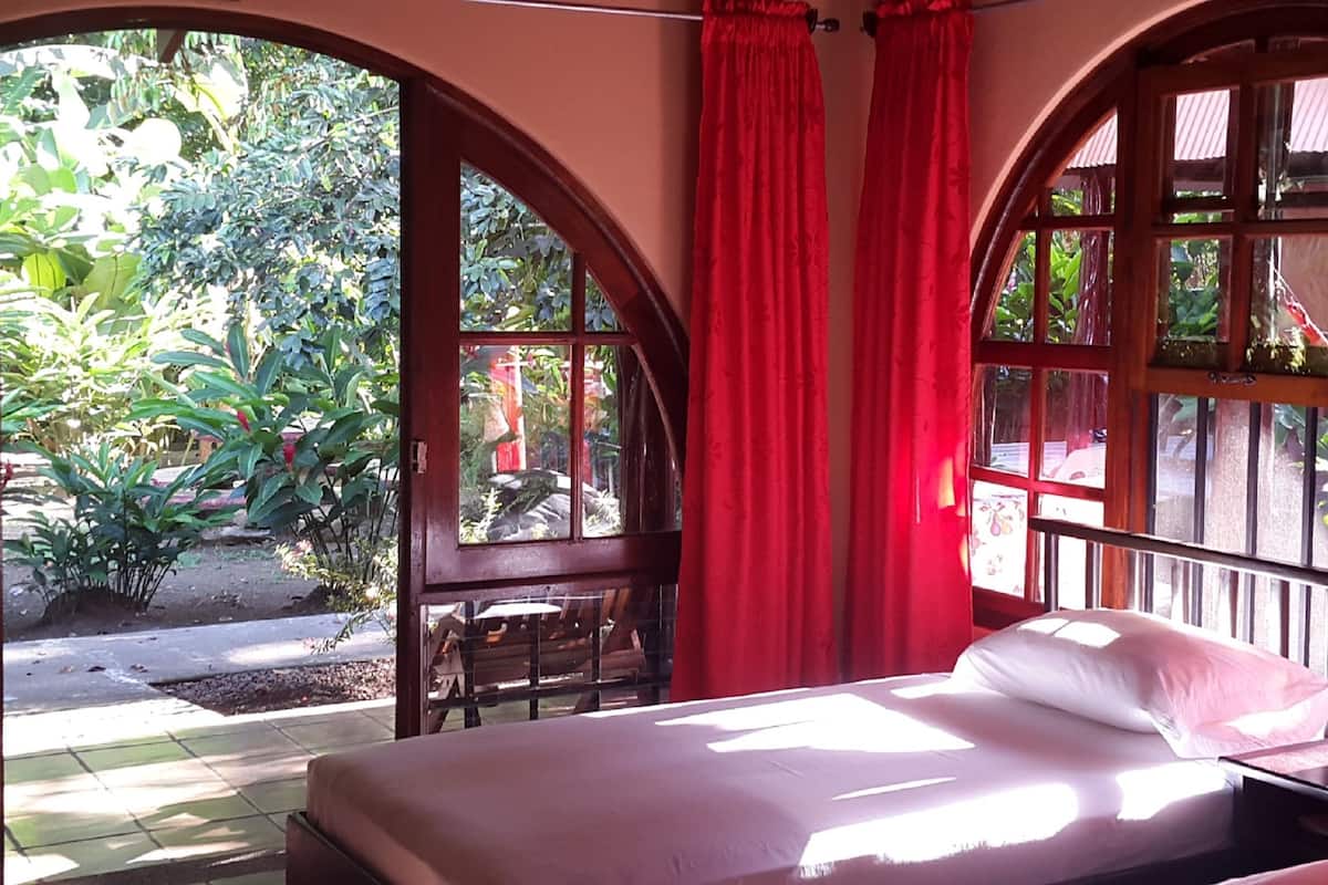 A room with a bed, a nightstand, and a window with red curtains. There is a view of greenery outside.