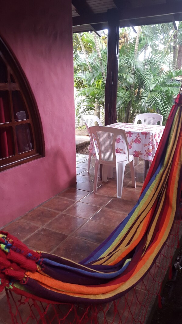 A hammock, a table with a floral tablecloth, and chairs outside a building with a pink wall and a window.
