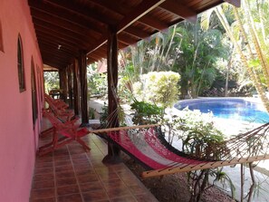 A covered patio with a pool, wooden chairs, and a hammock.