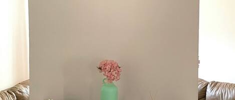A room with a dark wooden desk, a teal vase with pink flowers, a framed picture on the wall, and a brown leather couch.