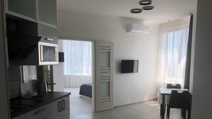 Una habitación de hotel moderna con kitchenette, un televisor de pantalla plana montado en la pared, un área de comedor con mesa y sillas, y un ventanal con cortinas translúcidas.