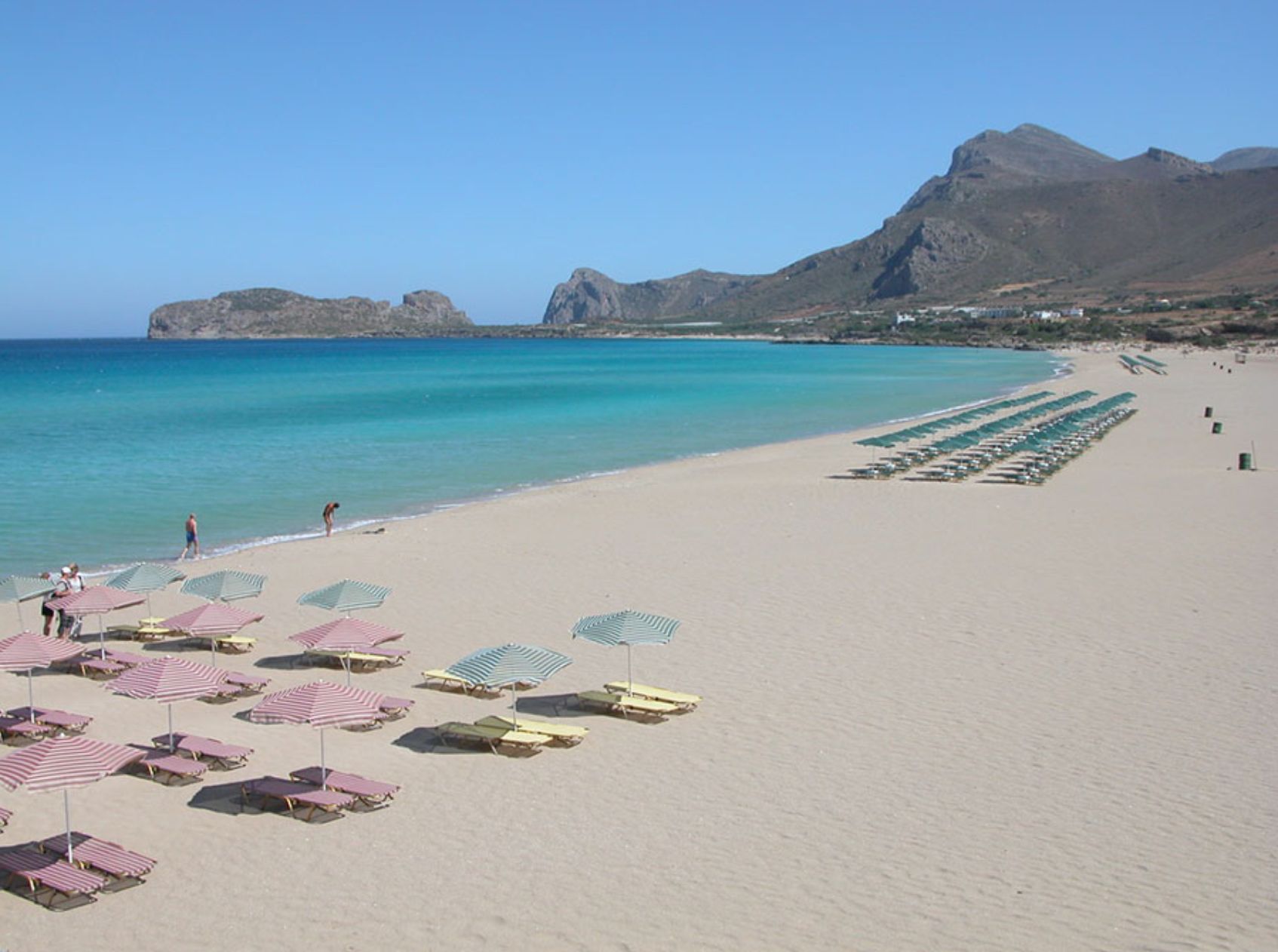 A beach with rows of sun loungers and umbrellas, a clear blue sea, and mountains in the background.
