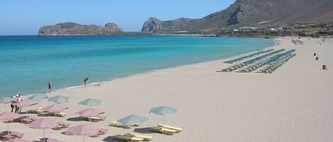A beach with rows of sun loungers and umbrellas, a clear blue sea, and mountains in the background.