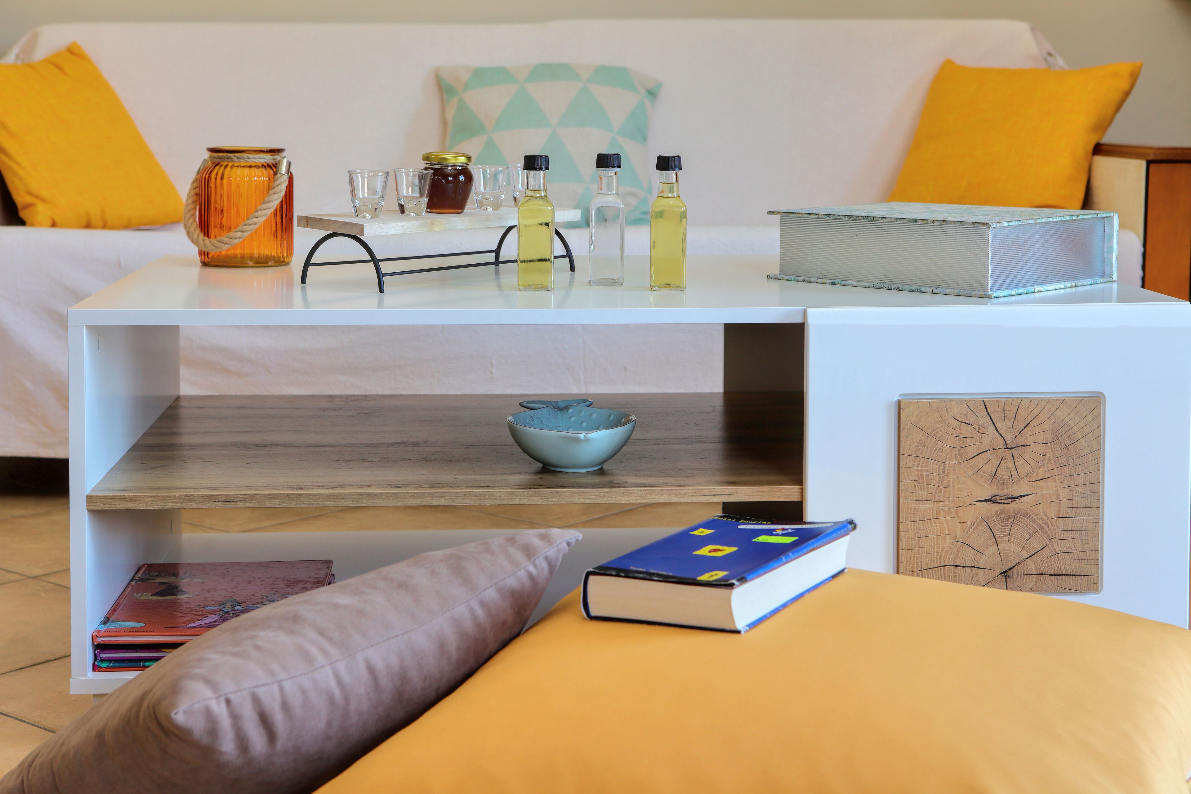 Habitación ordenada con una cama blanca, una mesita con varios objetos, un tazón azul y un libro sobre un cojín amarillo.