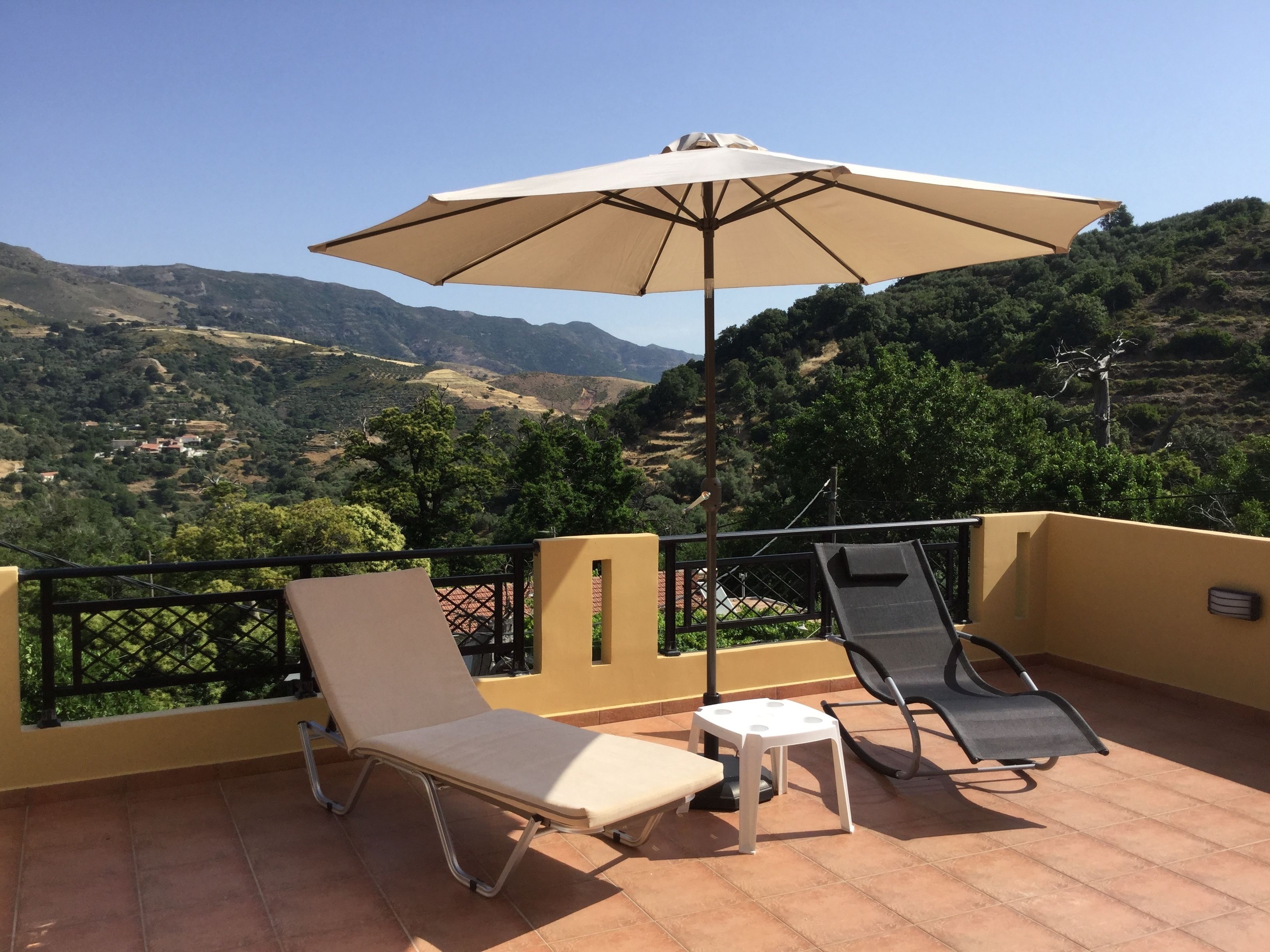 A terrace with two lounge chairs and a table, an umbrella, and a view of a hilly landscape.