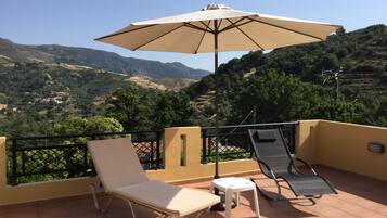 A terrace with two lounge chairs and a table, an umbrella, and a view of a hilly landscape.