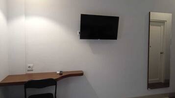 A room with a flat-screen TV mounted on the wall, a wooden desk, a black chair, and a door.