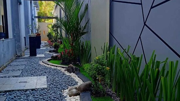 Un estrecho sendero de piedras y pequeñas plantas, flanqueado por muros con diseños geométricos y unidades de aire acondicionado.