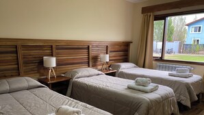 Quarto de hotel com três camas, cabeceira de madeira, abajures e vista para um prédio azul lá fora.