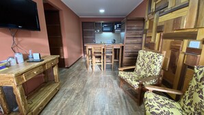Um espaço de estar compacto com mesa de madeira, sofá, TV fixada na parede e área de cozinha ao fundo.