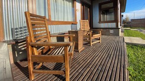 Um deck de madeira com duas cadeiras e uma mesa de madeira, uma parede de metal corrugado e uma janela com cortinas.